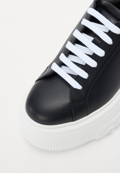 Black leather sneaker with white laces, rounded toe, and thick white rubber sole featuring textured accents and stitching details.