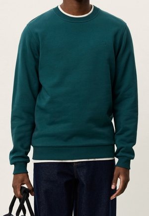 Mann trägt ein türkisfarbenes Sweatshirt über einem weißen Hemd, dunkle Jeans, hält eine schwarze Tasche mit beiden Händen an den Seiten.
