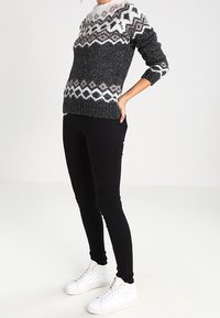 Maglione grigio lavorato a maglia con un motivo bianco e scuro sulla parte superiore, abbinato a jeans skinny neri e sneakers bianche, in piedi contro uno sfondo neutro.