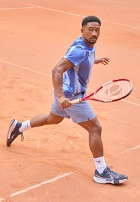 Athlet in einem hellblauen Mesh-T-Shirt mit kurzen Ärmeln und passenden Shorts, der auf einem Sandplatz läuft, während er einen roten Tennisschläger hält und dunkle Sneaker trägt.