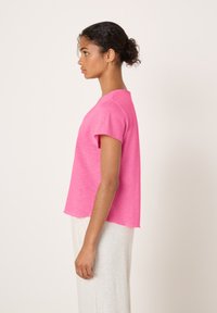 Ženska z nakodranimi lasmi v figi, oblečena v svetlo roza kratkoročno majico in svetle hlače, stoji v profil proti enostavni podlagi.