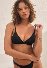 Top triangolo nero con spalline sottili, caratterizzato da una texture di tessuto liscia. La modella è in posa con le braccia incrociate, mettendo in evidenza il design.