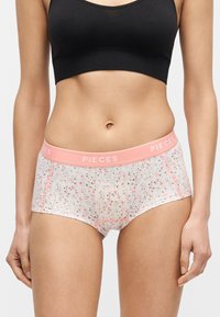 Γυναίκα που φοράει μαύρο κοντό τοπ και λευκά λουλουδάτα boyshort εσώρουχα με ροζ λάστιχο στη μέση με την ένδειξη "PIECES."