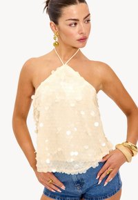 Femme portant un top halter crème avec de grandes paillettes, un short en denim, des boucles d'oreilles en forme de coquillage dorées, et de chunky bracelets au poignet droit.