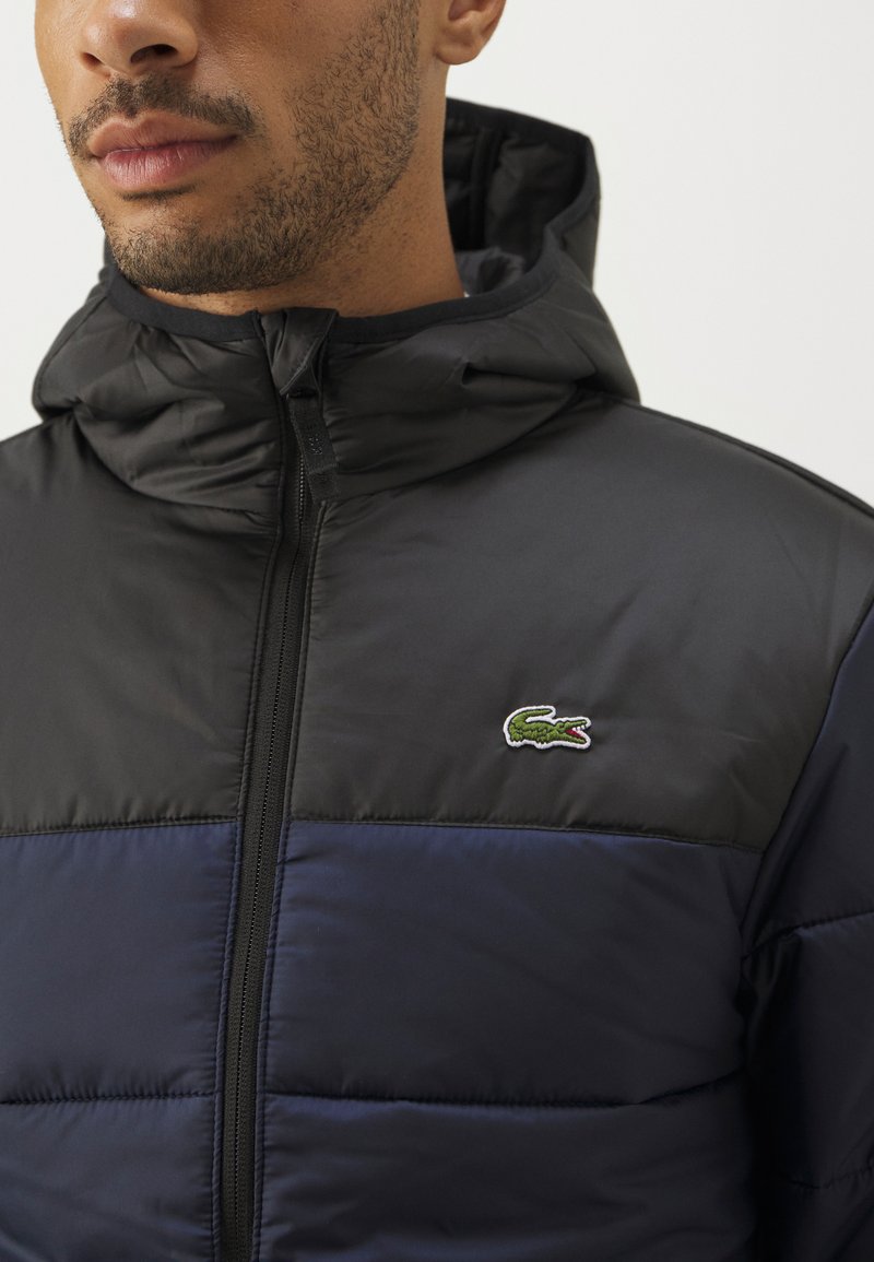 Hooded Pufferjacke mit schwarzem Oberteil und marineblauem Unterteil. Hergestellt aus glattem, glänzendem Stoff mit einem kleinen gestickten Logo.