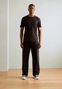 Sort kortærmet t-shirt parret med løstsiddende sorte jeans og sort-hvide sneakers, sat mod en neutral væg og trægulv.