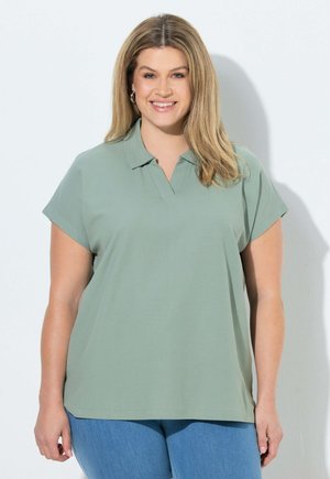 Uśmiechnięta kobieta o długich blond włosach, ubrana w jasnozieloną koszulkę polo z krótkim rękawem i niebieskie dżinsy, stojąca przy białej ścianie.