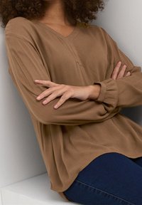 Bruine V-hals top met losse mouwen en een gestructureerde stof. Opgevouwen manchetten en een ontspannen pasvorm, gecombineerd met donkerblauwe jeans. Eenvoudig, modern ontwerp.