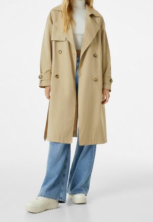 Kvinde iført beige trenchcoat over hvid kort rullekrave, blå vidde jeans og hvide platformsneakers på hvid baggrund.