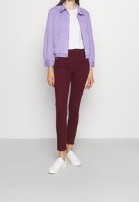 Chaqueta de mezclilla morada con cremallera, camiseta blanca, pantalones slim-fit burdeos y zapatillas blancas, exhibiendo un atuendo casual con vibrantes contrastes de color.