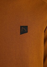 Tissu tricoté couleur rouille avec un petit patch carré noir présentant une ligne orange abstraite et inclinée cousue en son centre.