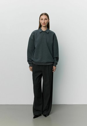 Une femme se tient contre un mur uni, portant un polo gris foncé oversize à manches longues, un pantalon large noir et des chaussures noires pointues.