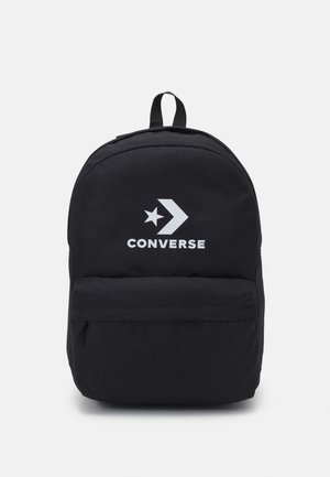 Mochila de tela negra con forma redondeada. Cuenta con un bolsillo frontal, un asa superior y un logo impreso en blanco con un diseño de estrella y flecha.