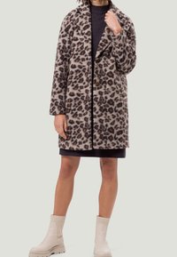 Manteau en imprimé léopard avec un col cranté, deux boutons, fabriqué en matériau doux. Porté par-dessus une robe sombre avec des bottines beige.