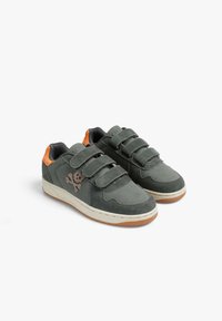Zapatillas grises para niños con tres tiras de velcro, detalles en naranja en el talón, logo de calavera y huesos cruzados, y suelas blancas con tracción naranja.