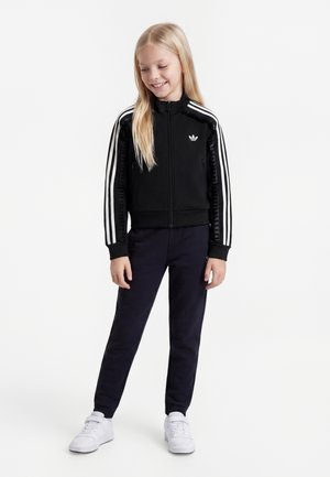 Pige med langt blondt hår iført en sort Adidas jakke med hvide striber og mørke bukser, der står og smiler, mens hun kigger ned.