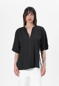 Haut noir à manches courtes avec un décolleté en V plongeant et une fermeture éclair à l'avant. Présente une coupe ample et des fentes sur les côtés, fabriqué dans un tissu lisse.