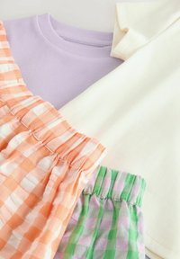Tre beklædningsgenstande: en lavendel ribbet T-shirt, en cremefarvet ribbet T-shirt, og to ternede shorts i orange og grøn/lilla nuancer, med elastisk talje.