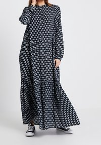 Kobieta w długiej czarnej sukience z białym wzorem liści i czarno-białych sneakersach, stojąca na jednolitym białym tle.