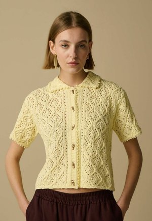 Femme portant un haut jaune en crochet à manches courtes avec col et boutons, et un pantalon marron, debout devant un fond beige.