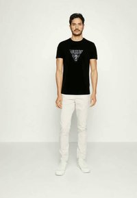Tricou negru din bumbac cu un design grafic triangular, pe care se află cuvântul "GUESS?" scris cu argintiu. Asortat cu blugi albi și sneakers albi.