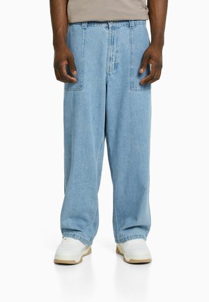 Jean en denim bleu clair à coupe ample avec de grandes poches plaquées à l'avant, porté avec des baskets blanches épaisses et un haut beige.