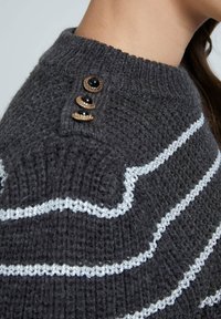 Gray knitted sweater with silver wavy stripes, featuring three decorative buttons at the shoulder and a ribbed neckline.