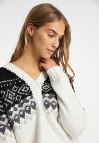 Cardigan bianco con motivi geometrici neri e grigi, dotato di scollo a V, due bottoni e polsini a costine. Texture in morbido tessuto a maglia.