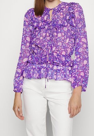 Kobieta ubrana w fioletową, kwiecistą bluzkę z falbanami, długimi rękawami i dekoltem typu keyhole, w połączeniu z białymi spodniami.