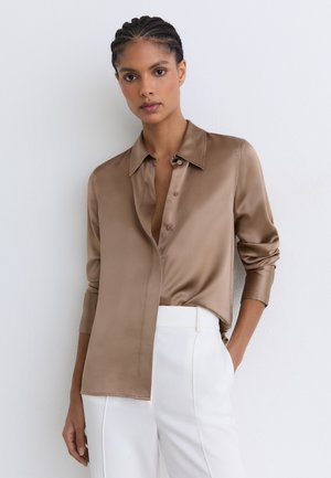 Donna che indossa una camicia di raso marrone lucida con bottoni e pantaloni bianchi a vita alta, in piedi con una mano in tasca contro un muro bianco.