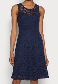 Marinblå ärmlös spetsklänning med blommönster, figursydd överdel och utsvängd kjol, med en ellypsformad spetskrage och textur.