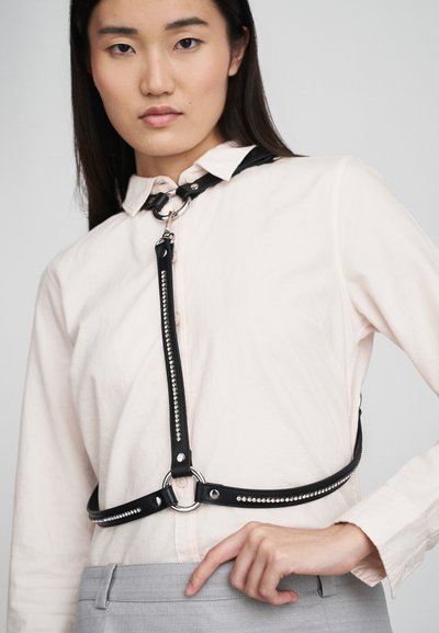 Čierny kožený postroj so striebornými cvokmi, s nastaviteľnými popruhmi a centrálnym krúžkom, skombinovaný s svetlým tričkom a šedými nohavicami.