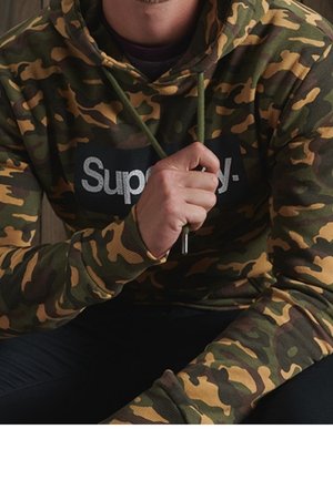 Homme portant un sweat à capuche camouflage vert et marron avec le texte « Supply. », tenant le cordon de la capuche près du menton.