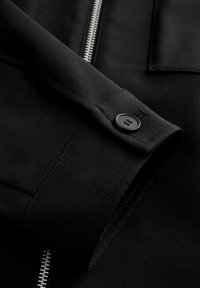 Veste noire avec une texture de tissu lisse, fermeture éclair argentée et un détail de bouton sur la manche, mettant en avant des lignes épurées et un design minimaliste.