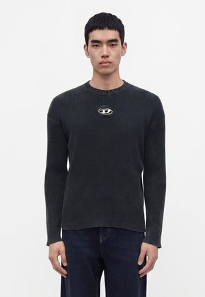 Giovane uomo che indossa un maglione a coste scuro con un taglio ovale unico e un emblema metallico sul petto, abbinato a jeans blu scuro.