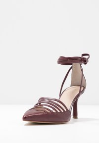 Scarpa con tacco alto bordeaux, punta affusolata, con più cinturini orizzontali sulla parte superiore e dettaglio di lacci alla caviglia. Texture in pelle liscia.