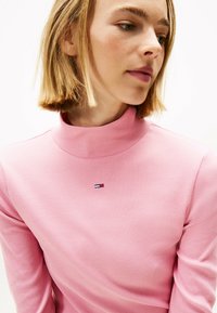 Hellrosa gerippter Rollkragenpullover mit langen Ärmeln, der ein kleines, buntes Logo auf der Brust trägt. Glatte Textur, figurnaher Schnitt.