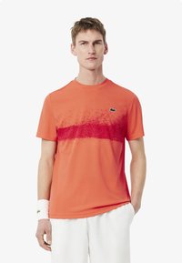 Orange idrottströja med en gradient röd sprutdesign över bröstet, rundad halsringning och Lacoste-logotyp på vänster sida.