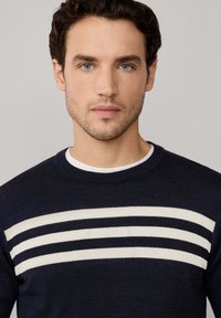 Maglione lavorato a maglia blu navy con strisce orizzontali color crema sul petto, dotato di scollo rotondo e una texture liscia.