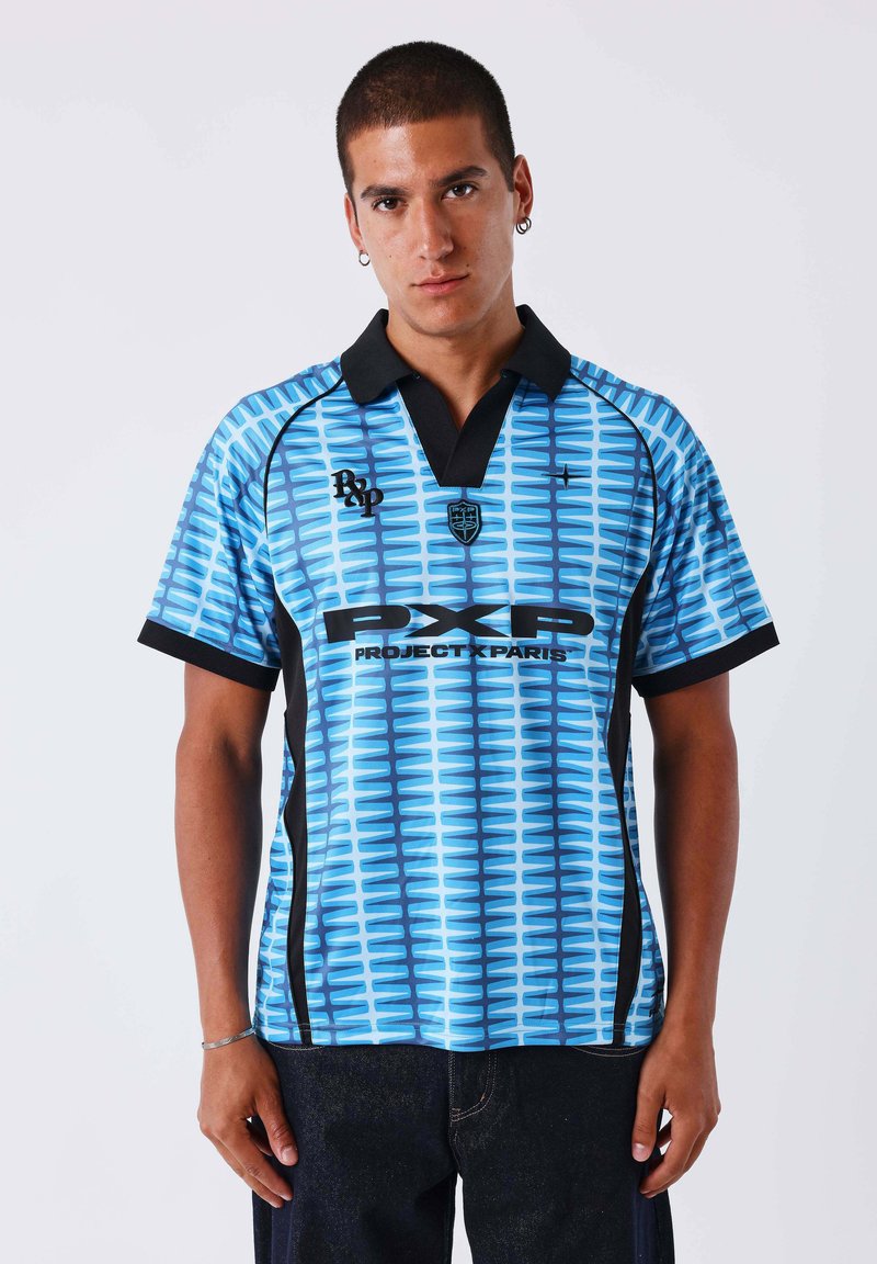 Homme portant un polo à manches courtes bleu à motifs avec col noir et texte « PX Paris », debout devant un fond clair uni.