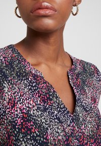 Navy-blaues Bluse mit V-Ausschnitt, aus weichem Stoff mit einem bunten, gesprenkelten Muster in Pink, Weiß und Grün. Goldene Creolen sind sichtbar.