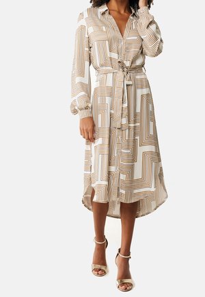 Femme portant une robe midi à manches longues ceinturée beige et blanche à motifs géométriques, avec des boutons sur le devant et des talons métalliques à bride cheville.