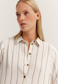 Weiß gestreifte Bluse mit braunen vertikalen Streifen, Knopfleiste, und braunen Knöpfen. Hergestellt aus leichtem Stoff mit weicher Textur.