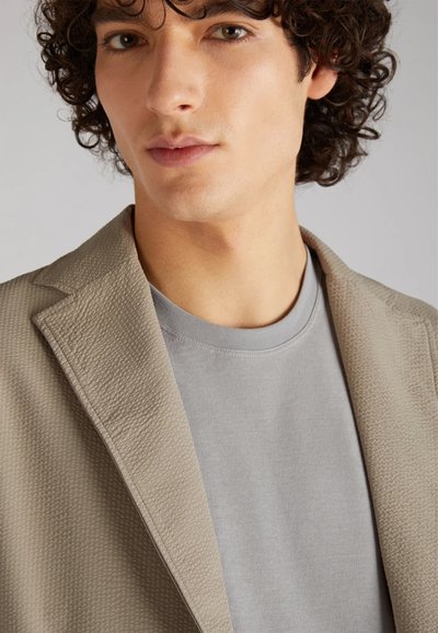 Blazer beige testurizzato sopra una t-shirt grigia chiara. Il blazer presenta un rever a punte e una superficie con un motivo, creando interesse visivo.