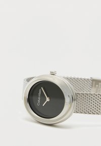 Ρολόι με μαύρο στρογγυλό καντράν, ασημένια μεταλλική θήκη και μεταλλικό λουράκι τύπου πλέγματος. Διαθέτει σήμα Calvin Klein και απλά δείκτες για τις ώρες και τα λεπτά.