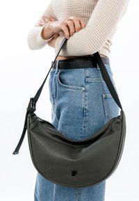 Person wearing blue jeans and a beige knit sweater holding a black crescent-shaped leather shoulder bag with a zipper.