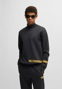 Svart sweatshirt med hög krage, prydd med ett guld horisontellt rand och "BOSS"-logotypen på den nedre framsidan. Tillverkad av ett slätt, strukturerat tyg.