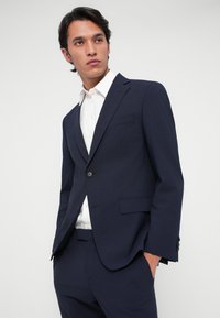 Μαρινί κοστούμι με κομψή εφαρμογή, κλείσιμο με ένα κουμπί, καμπυλωτά υφάσματα και υφή, συνδυασμένο με λευκό πουκάμισο.