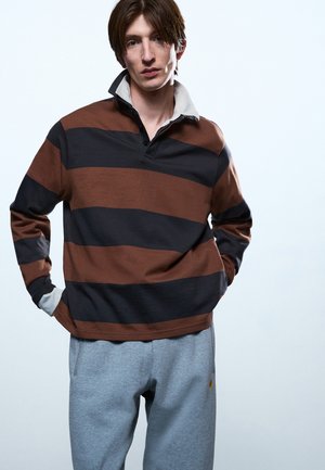 Νέος άνδρας που φοράει ένα καφέ και μαύρο ριγέ rugby πουκάμισο με όρθιο γιακά και ανοιχτό γκρι φόρμα, στέκεται με τα χέρια στις τσέπες.