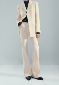 Personne portant un blazer crème, un pantalon large crème, un col roulé noir, une ceinture noire et des chaussures pointues noires, debout devant un fond bleu pâle.